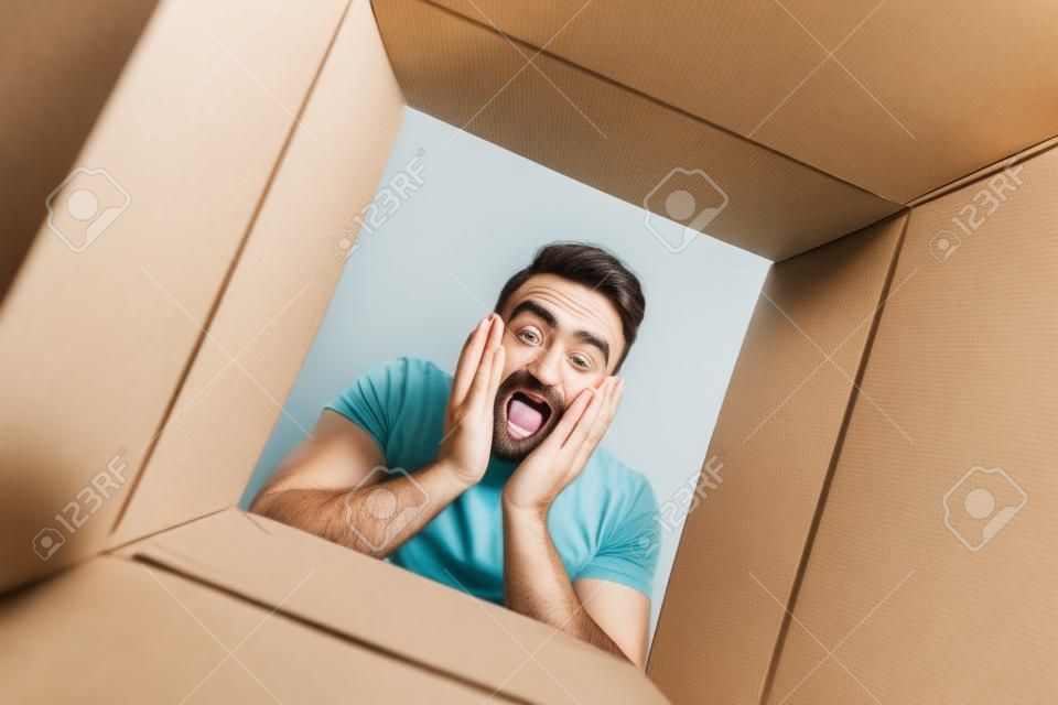 Der überraschte Mann packt aus, öffnet Karton und schaut hinein. Das Paket, die Lieferung, die Überraschung, das Geschenk-Lifestyle-Konzept. Konzepte für menschliche Emotionen und Gesichtsausdrücke
