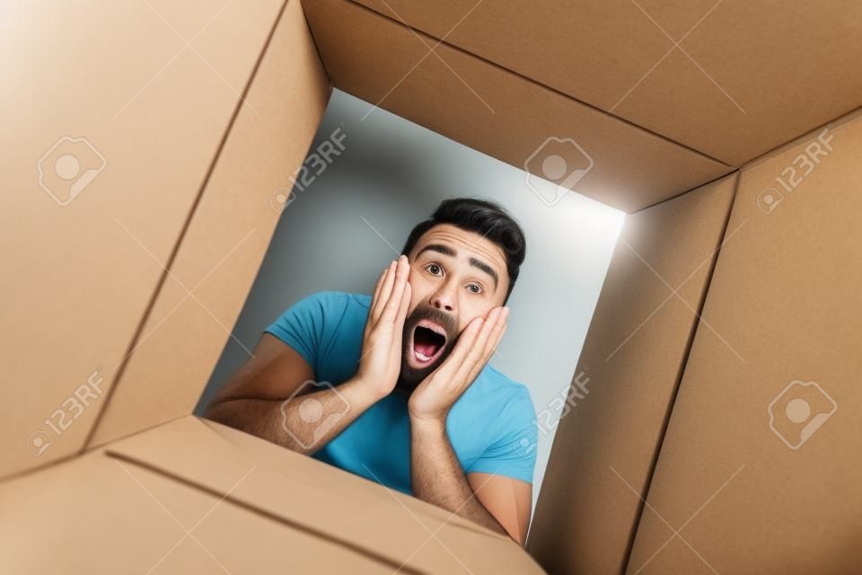 Zaskoczony mężczyzna rozpakowujący otwierające się pudełko kartonowe i zaglądający do wnętrza przesyłki, prezent-niespodzianka, koncepcja stylu życia, ludzkie emocje i mimika