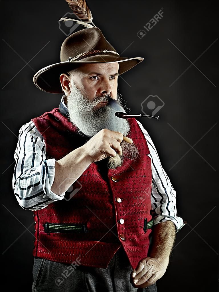 dramatic portrait of gray bearded senior man in hat smoking tobacco pipe. view of Austrian, Tyrolean, Bavarian old man in national traditional costume in retro style.