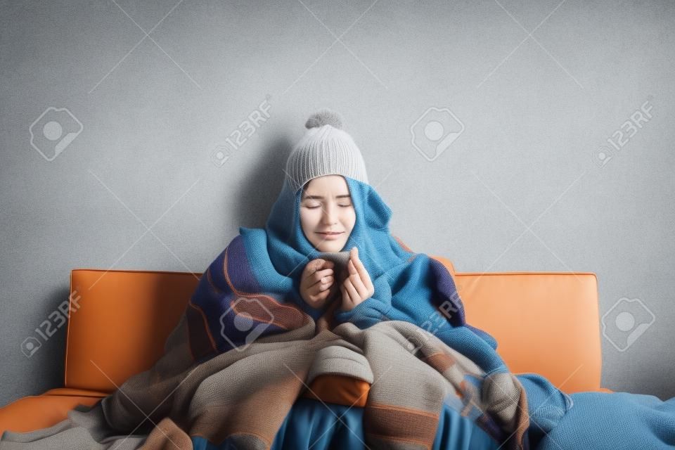 Młoda chora kobieta z grypą siedzi na kanapie w domu lub w studio, przykryta ciepłymi ubraniami z dzianiny. Choroba, grypa, koncepcja bólu. Relaks w domu. Koncepcje opieki zdrowotnej.