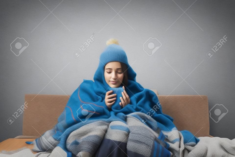 Młoda chora kobieta z grypą siedzi na kanapie w domu lub w studio, przykryta ciepłymi ubraniami z dzianiny. Choroba, grypa, koncepcja bólu. Relaks w domu. Koncepcje opieki zdrowotnej.