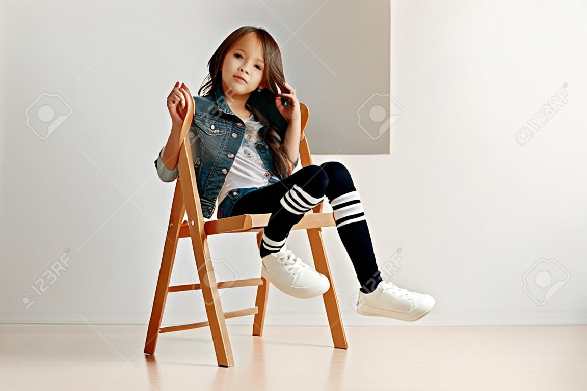Retrato completo da menina pequena bonito na roupa elegante do jeans que olha a câmera e que sorri contra a parede branca do estúdio.