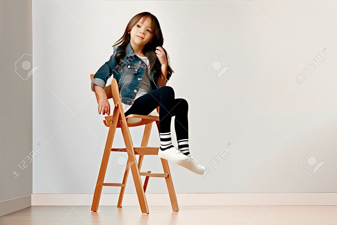 Retrato completo da menina pequena bonito na roupa elegante do jeans que olha a câmera e que sorri contra a parede branca do estúdio.