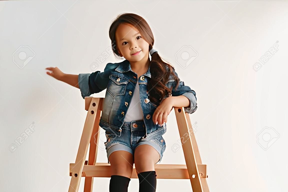 Das Porträt des süßen kleinen Mädchens in stilvoller Jeanskleidung, das die Kamera anschaut und lächelt und an der weißen Studiowand sitzt Konzept für Kindermode