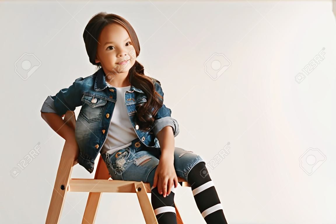 Das Porträt des süßen kleinen Mädchens in stilvoller Jeanskleidung, das die Kamera anschaut und lächelt und an der weißen Studiowand sitzt Konzept für Kindermode
