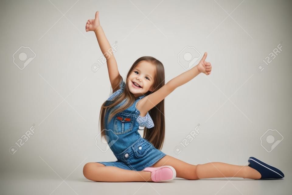 Pełna długość portret słodkie małe dziecko dziewczynka w ubrania stylowe dżinsy patrząc na kamery i uśmiechając się, stojąc na ścianie biały studio. Koncepcja mody dla dzieci