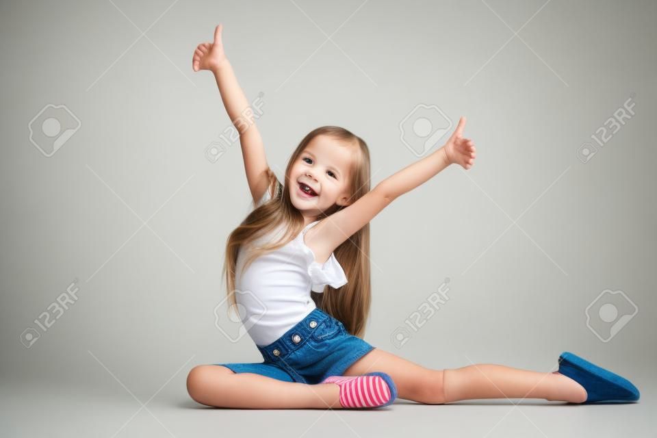 Ganzaufnahme eines süßen kleinen Mädchens in stilvoller Jeanskleidung, das die Kamera anschaut und lächelt und gegen die weiße Studiowand steht. Konzept für Kindermode