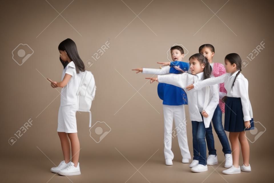 Little girl standing alone and suffering an act of bullying while children mocking in the background. Sad young schoolgirl standing on studio against white background.