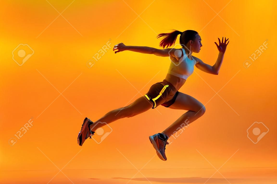 Speed. Dynamic image of young girl, professional athlete, runner in motion, training over orange studio background in neon light. Concept of sport. health and strength