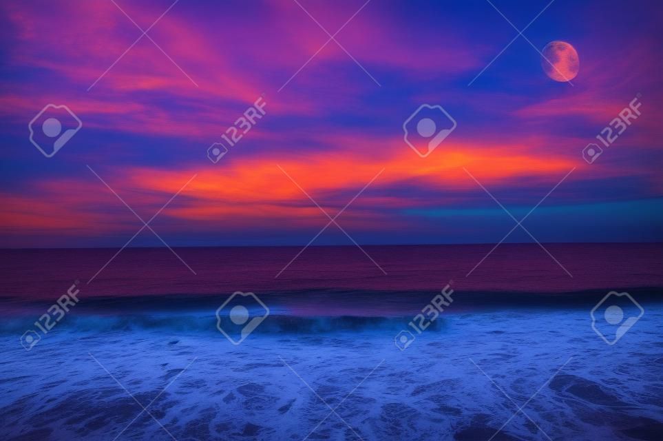 Ocean sunset moon is a colorful cloud filled sky over the ocean with a three quarter moon rising high in the sky.