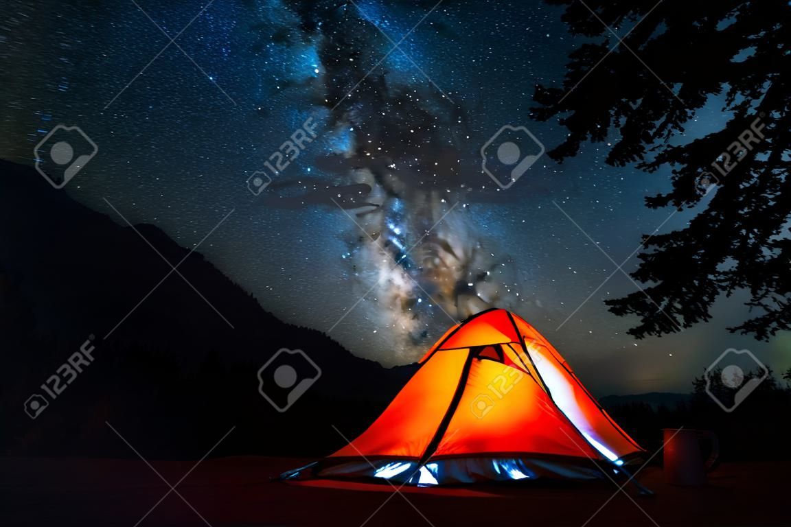 Namiot i nocne niebo. Podświetlony pomarańczowy namiot turystyczny i głębokie rozgwieżdżone niebo z drzewami na pierwszym planie.