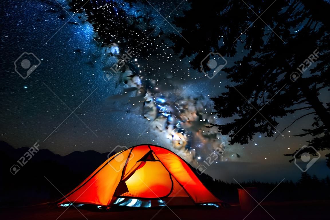 帳篷和夜空。被突出的橙色遠足的帳篷和深滿天星斗的天空與樹在前景。