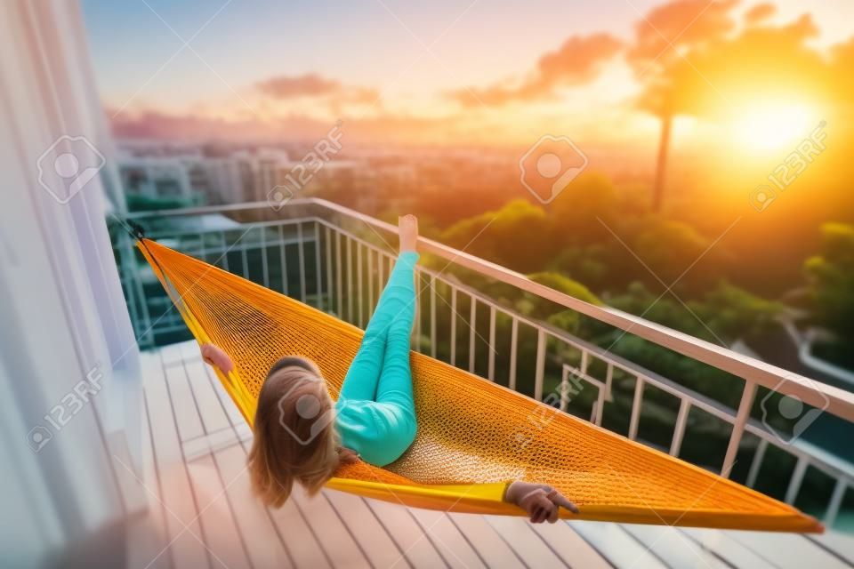 Woman relaxes in the hammock set on a balcony and enjoys sunset and tropical garden view. Tilt shift effect applied
