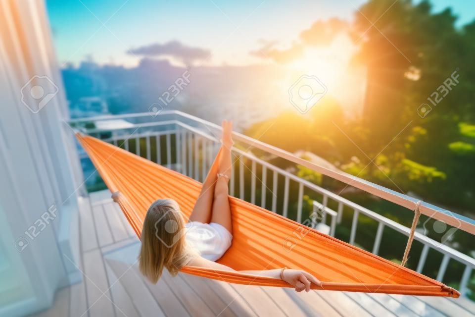 Woman relaxes in the hammock set on a balcony and enjoys sunset and tropical garden view. Tilt shift effect applied