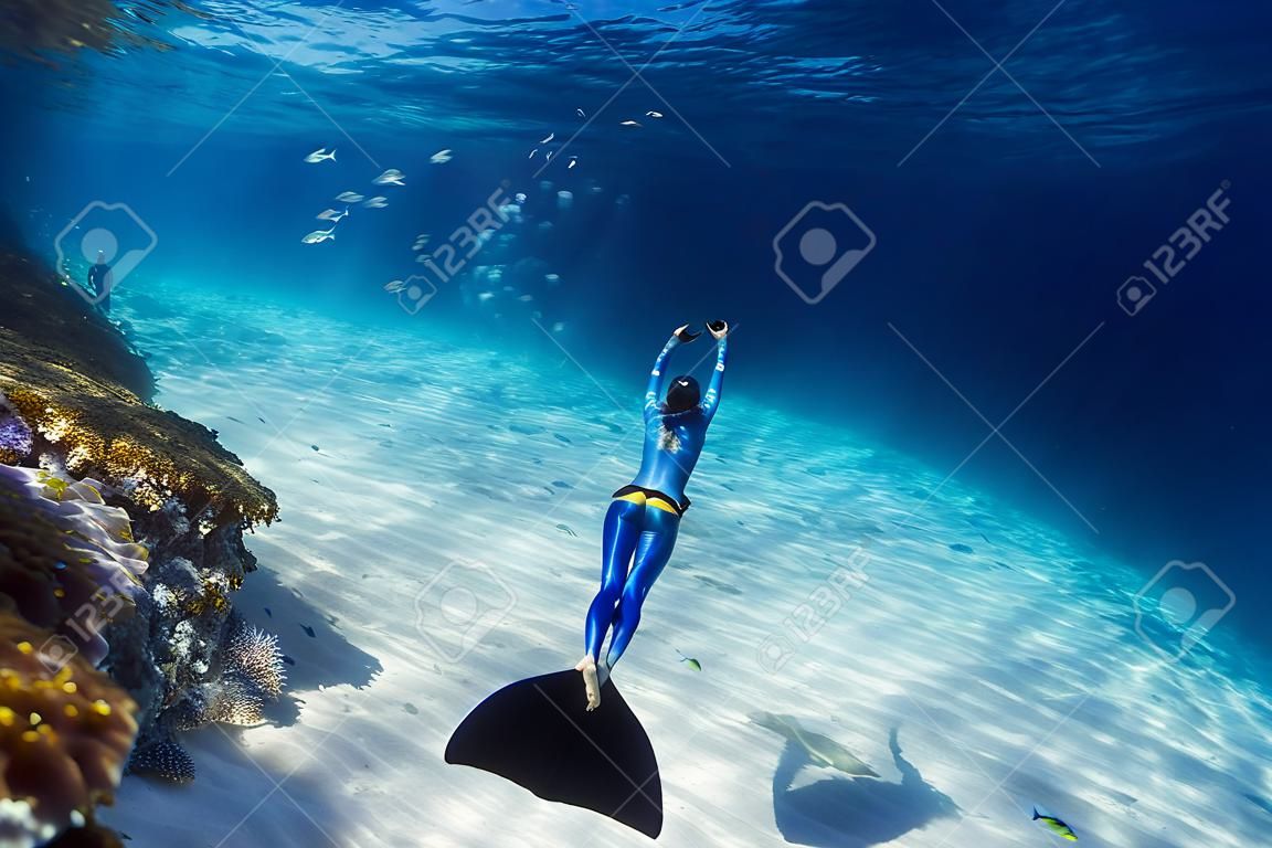 Freitaucherin in Monoflosse gleitet im Meer über den sandigen Boden mit Fischschwarm und passiert buntes Korallenriff