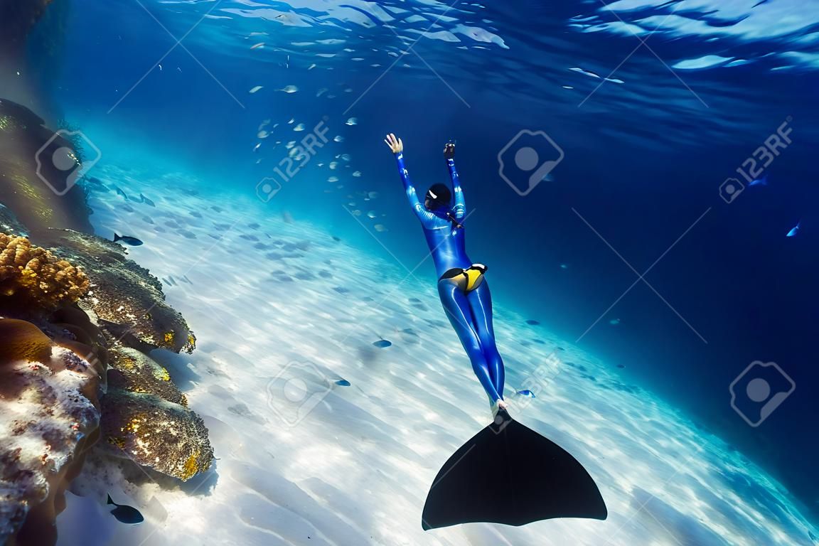 Freitaucherin in Monoflosse gleitet im Meer über den sandigen Boden mit Fischschwarm und passiert buntes Korallenriff