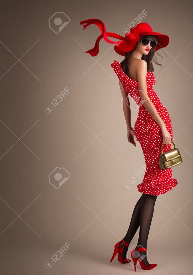 Стоя красивая молодая женщина, одетая в красную шляпу и горошек платье