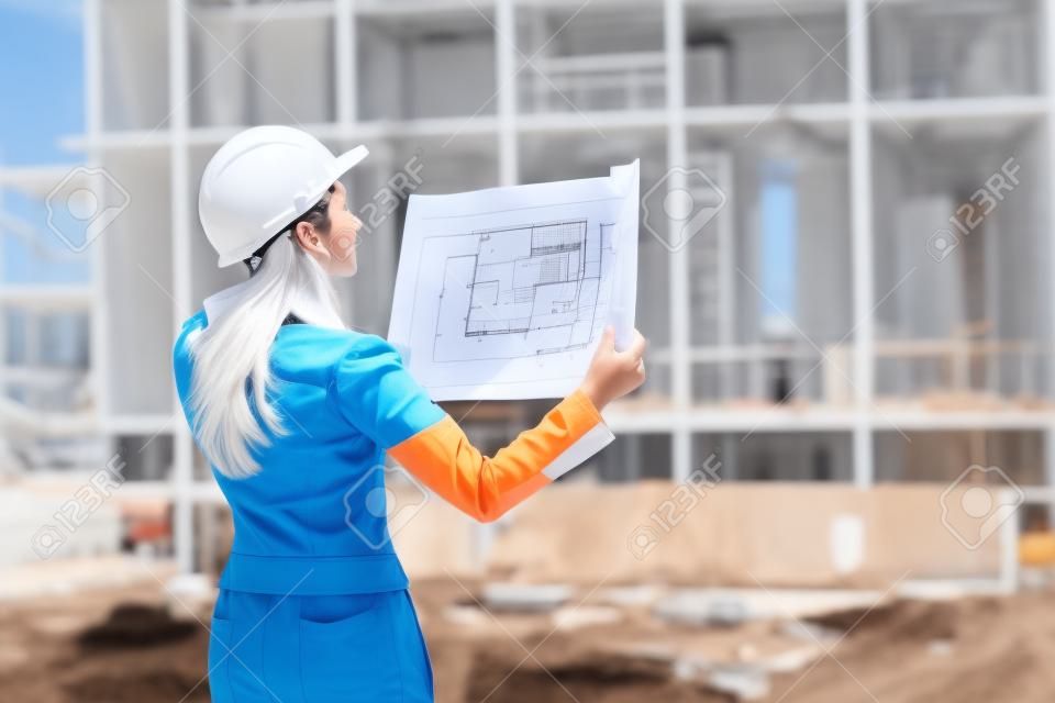 Successful young female engineer with white helmet checked new project and analyzing blueprints in front of construction site.