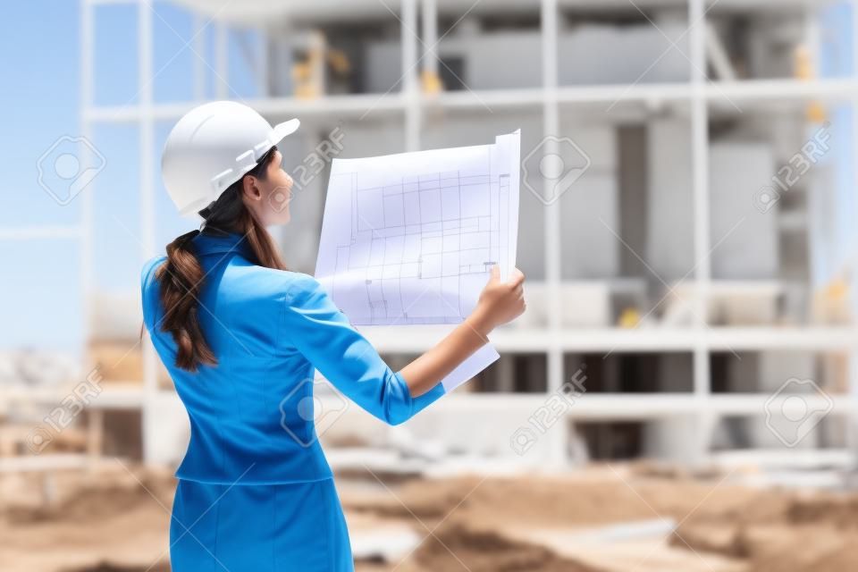 Successful young female engineer with white helmet checked new project and analyzing blueprints in front of construction site.