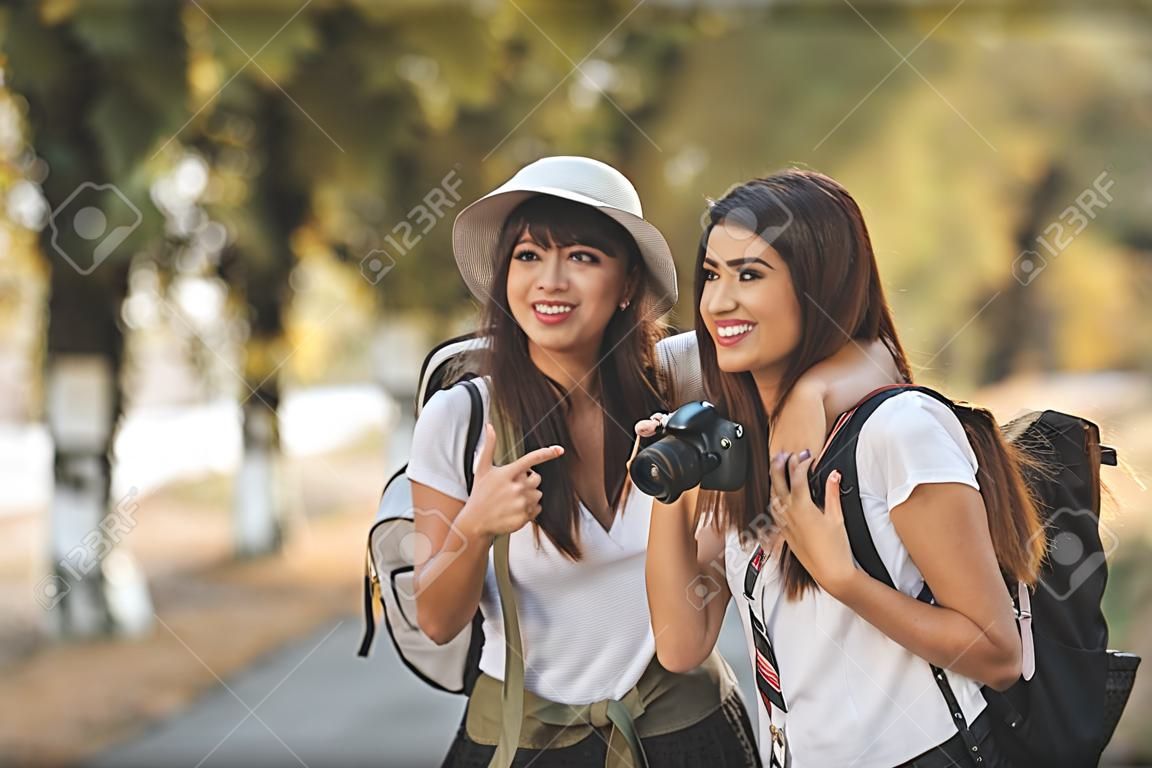 Two beautiful young smiling women with backpacks on their back are walking along the autumn sunny avenue and talking each other and pointing something.