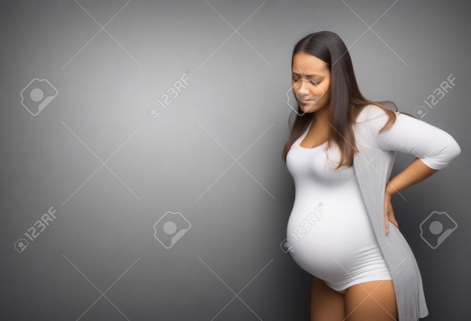 Mulher grávida com contrações, de pé no fundo cinzento do estúdio, tocando suas costas, espaço da cópia. Senhora expectante que sofre da dor durante o trabalho de parto, espaço da cópia