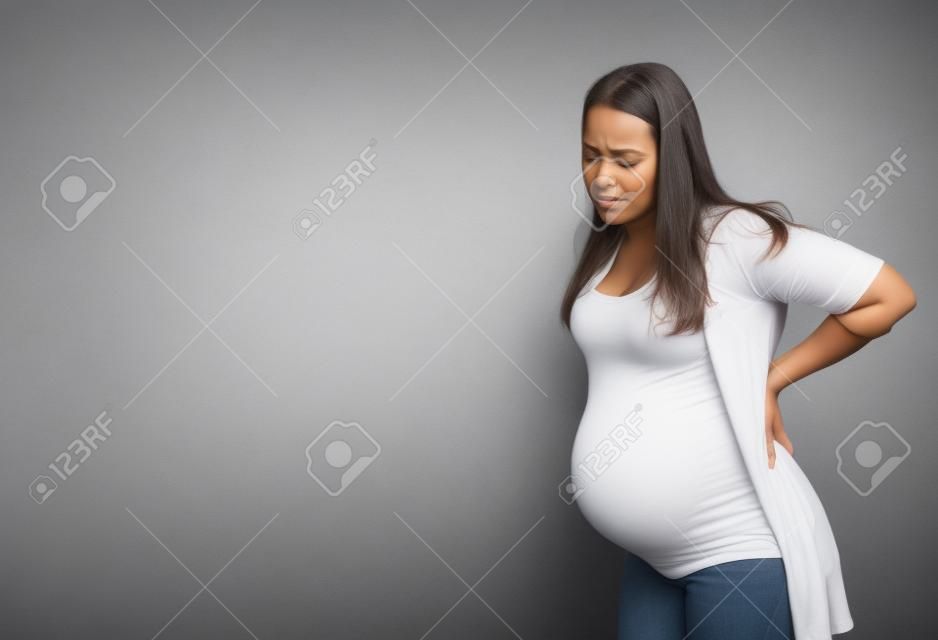 Pregnant woman with contractions, standing at gray studio background, touching her back, copy space. Expectant lady suffering from pain during labor, copy space