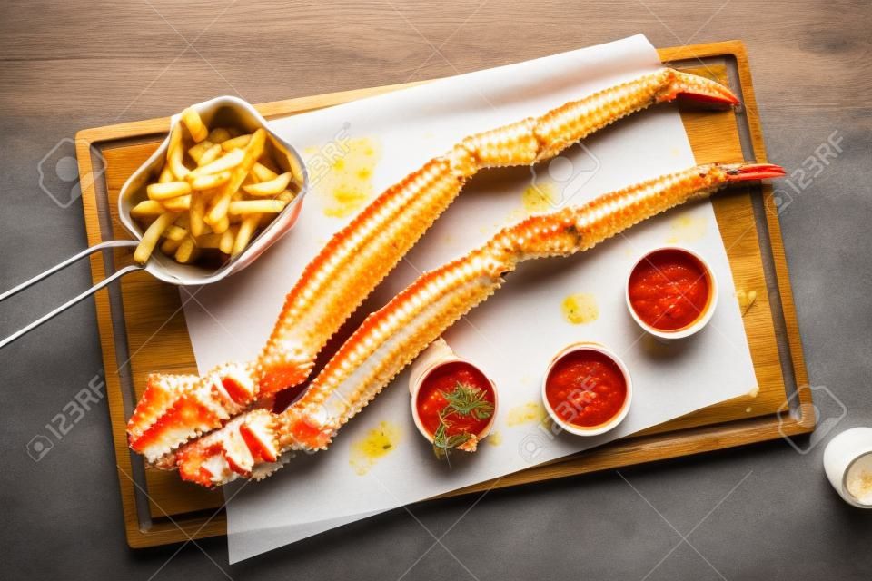 Alaskan king crab legs with butter and tomato sauces and french fries served on wooden board, top view