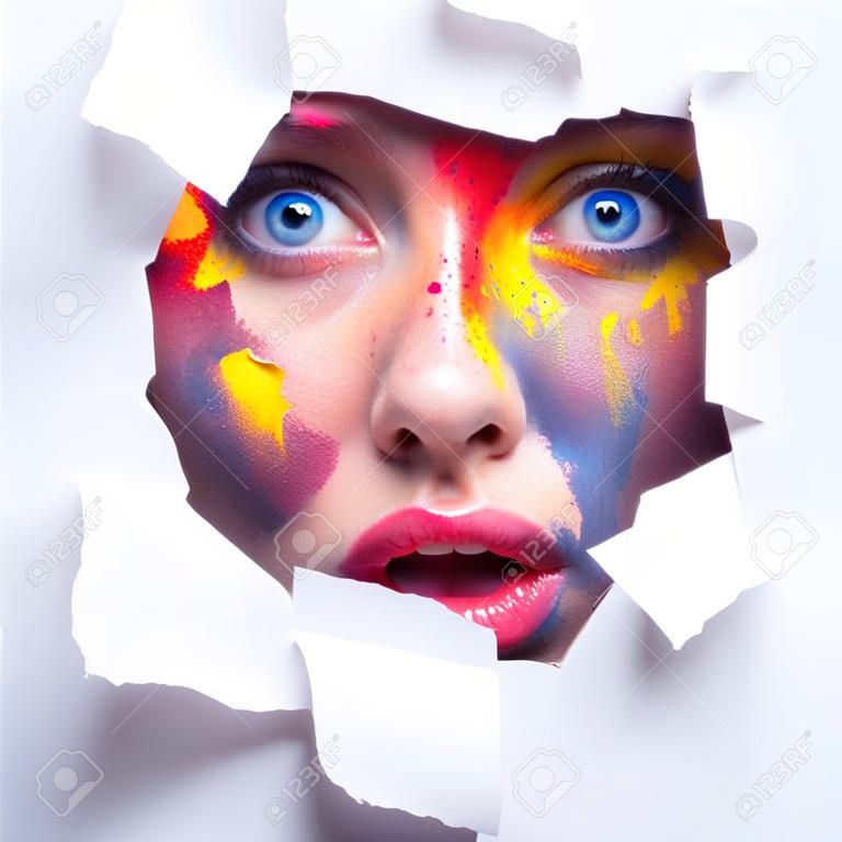 Necesita más color. Mujer conmocionada emocional con maquillaje creativo brillante asoma a través del agujero en papel blanco, espacio libre.