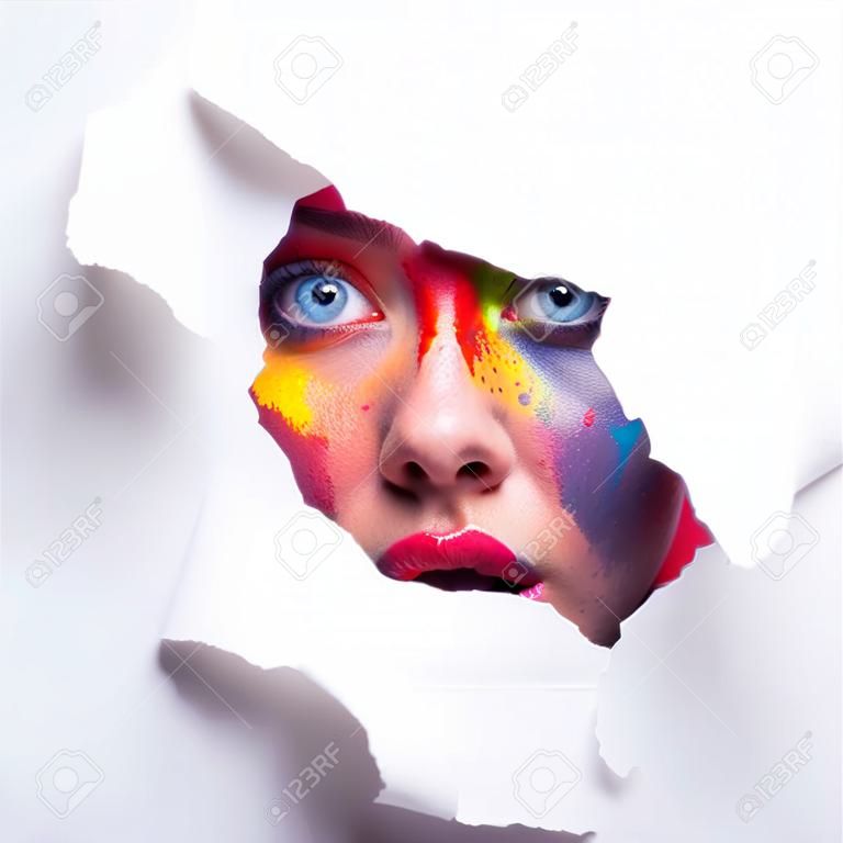 Need more color. Emotional shocked woman with bright creative makeup peeking through hole in white paper, free space.