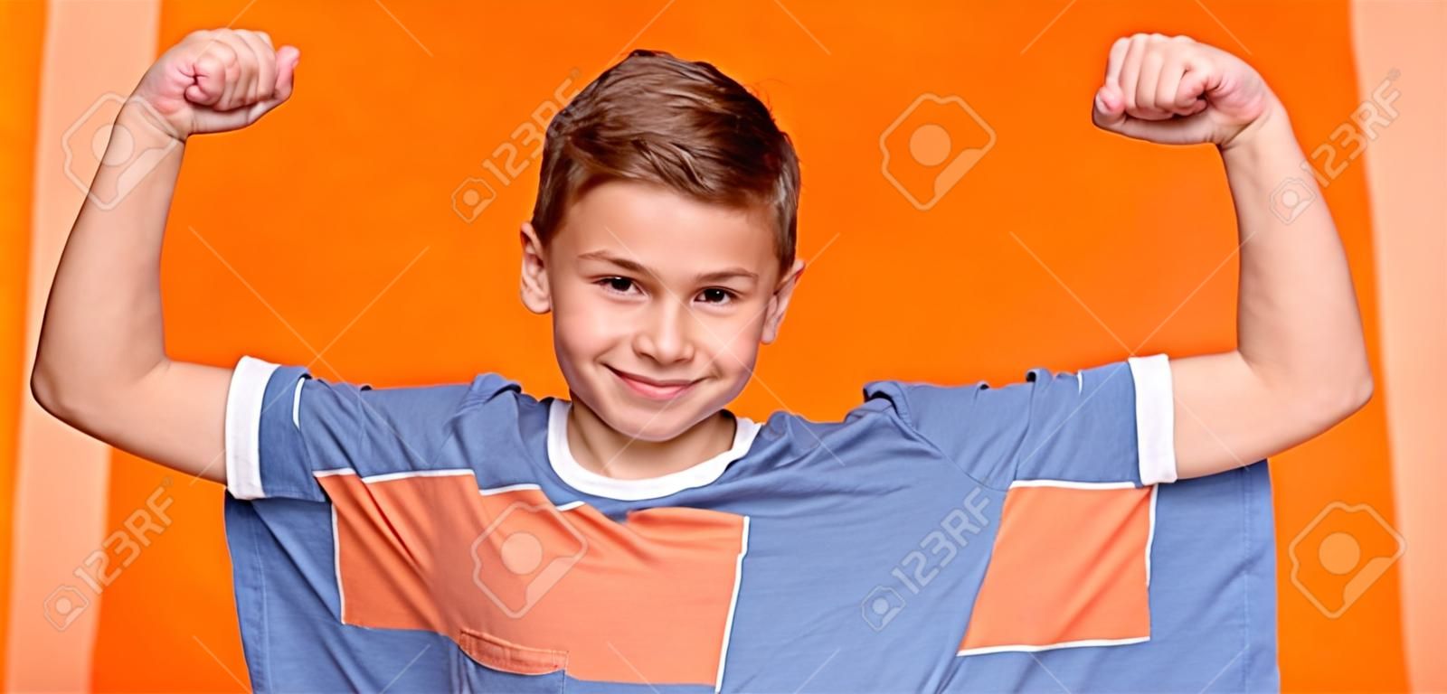 Starker kleiner Mann. Lächelnder Junge, der seinen Bizeps und seine Muskeln demonstriert, oranger Panoramahintergrund
