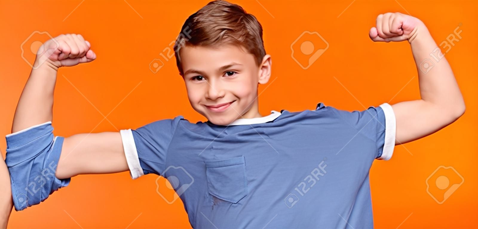 Starker kleiner Mann. Lächelnder Junge, der seinen Bizeps und seine Muskeln demonstriert, oranger Panoramahintergrund