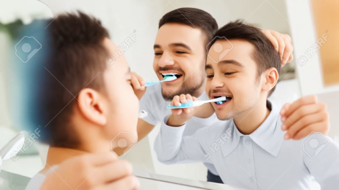 Little Daughter And Father Brushing Teeth Together In The Morning, Looking At Mirror