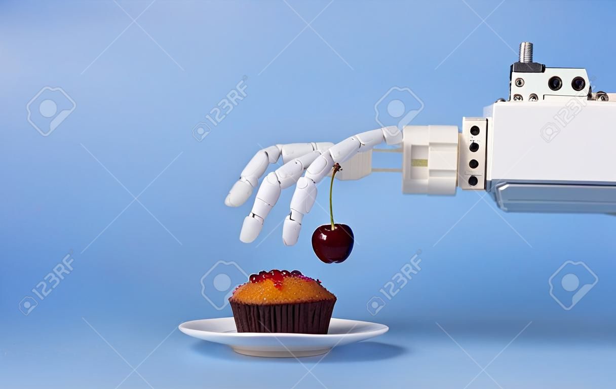 Robotyzacja kuchni. Ręka robota kładąca świeżą wiśnię na wierzchu babeczki, niebieskie tło