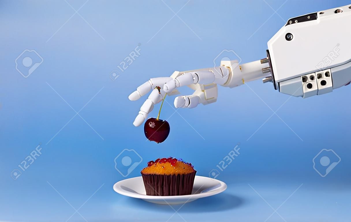 Robotización de cocinas. Mano de robot poniendo cereza fresca encima de la magdalena, fondo azul