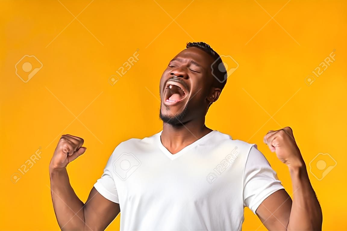 Happy Black Man Screaming In Delight And Shaking Fists Standing Over Yellow Studio Background.
