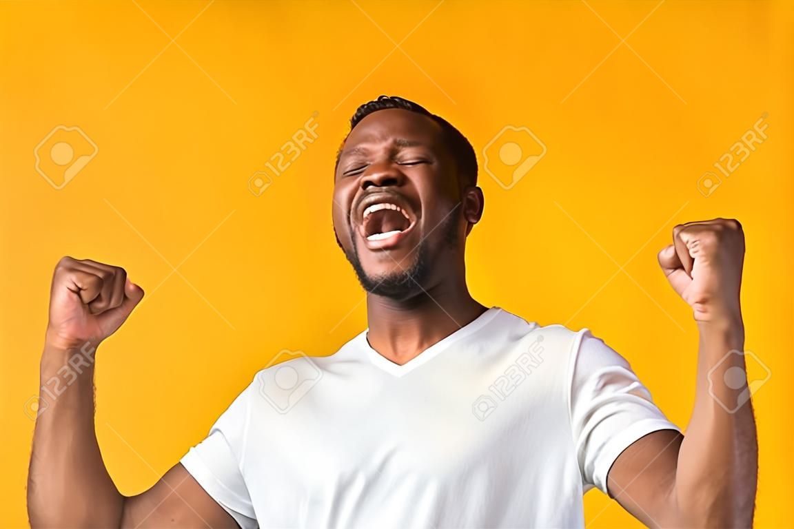 Hombre negro feliz gritando de alegría y sacudiendo los puños de pie sobre fondo de estudio amarillo.