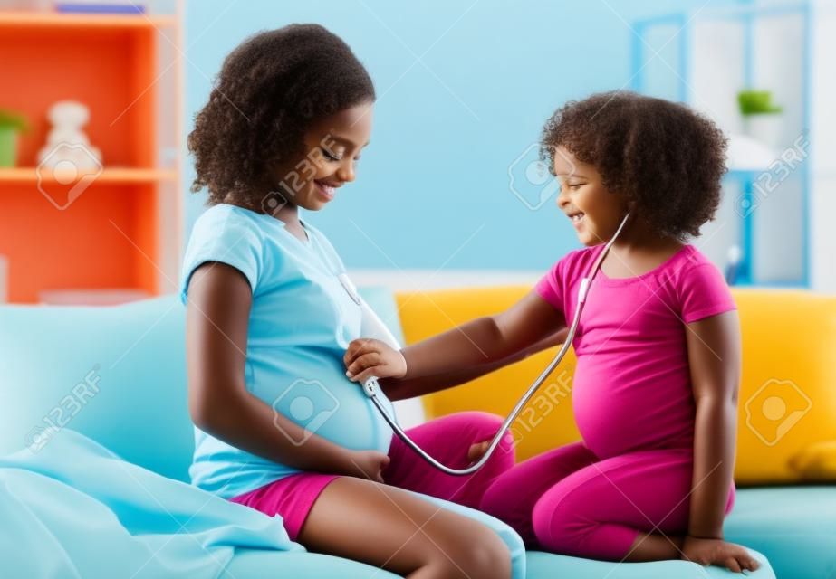 Neues Leben entdecken. Kleines schwarzes Mädchen, das den Bauch schwangerer Mütter mit Stethoskop hört, Arzt zu Hause spielt, freier Raum.