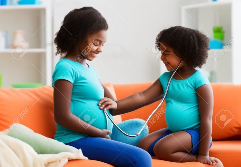 Neues Leben entdecken. Kleines schwarzes Mädchen, das den Bauch schwangerer Mütter mit Stethoskop hört, Arzt zu Hause spielt, freier Raum.