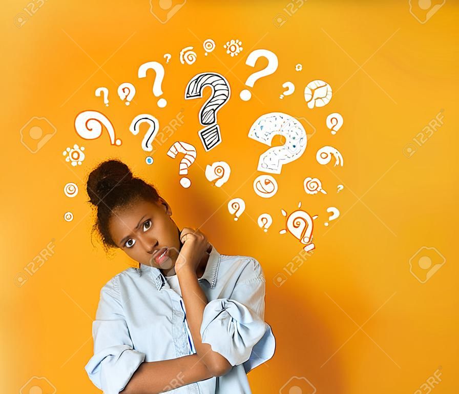 Perplejo pensativo joven adolescente afroamericana en camiseta casual posando sobre fondo naranja con signos de interrogación blancos