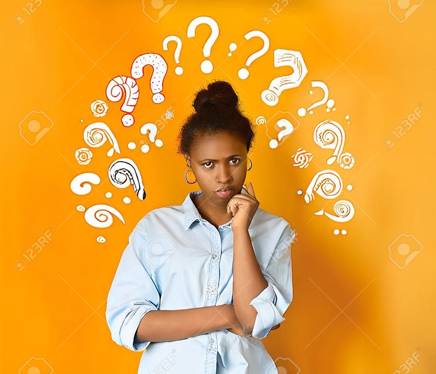 Perplejo pensativo joven adolescente afroamericana en camiseta casual posando sobre fondo naranja con signos de interrogación blancos