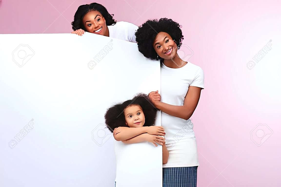 Family Offer. Happy African American Mother And Preschool Daughter Peeking Out Of Blank White Board With Free Space For Your Ad Over Pink Background