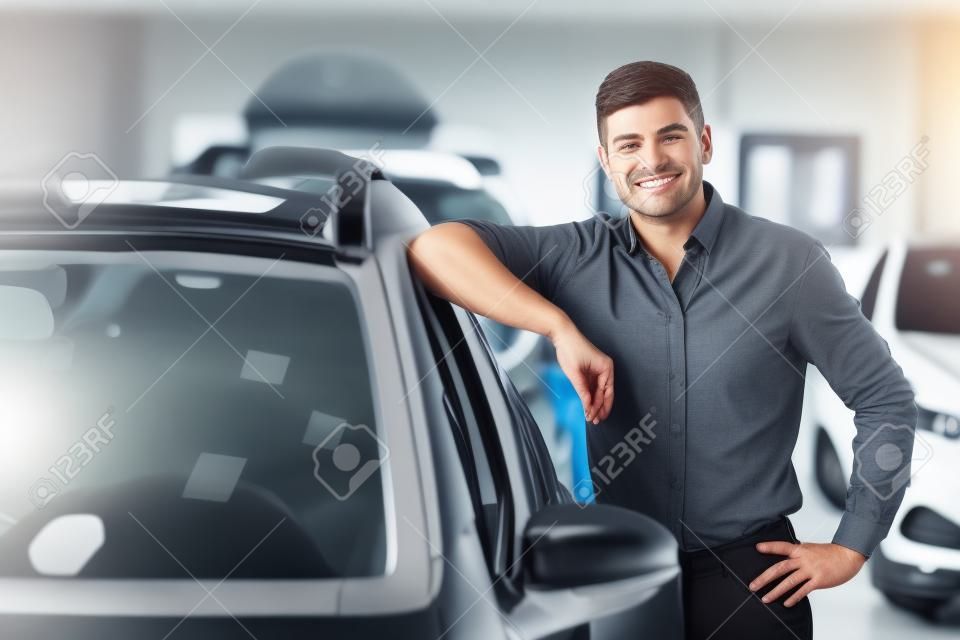 Pojęcie własności. Przystojny mężczyzna oparty o swój nowy samochód stojący w centrum dealerskim, wolna przestrzeń