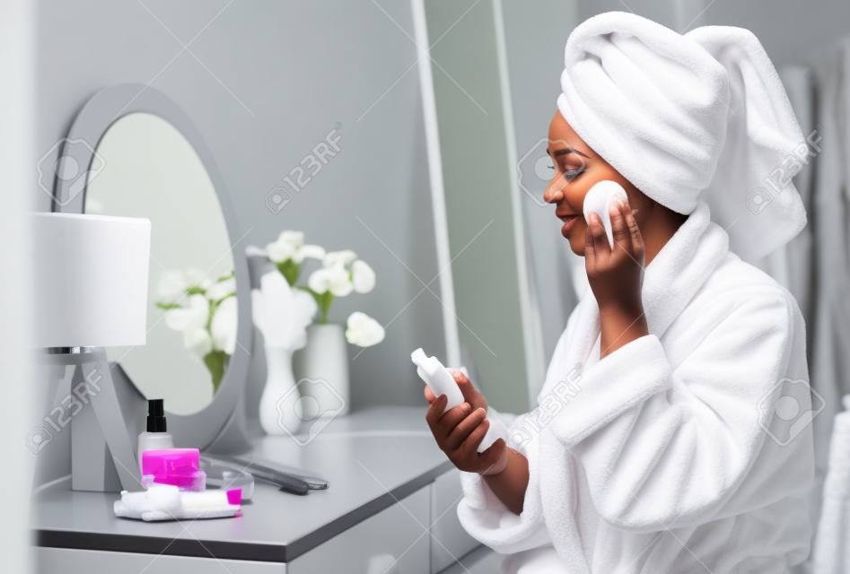 Mooie zwarte vrouw schoonmaken gezicht van make-up met reinigende melk en katoenen pad thuis, verpakt in badjas en handdoek, zittend aan dressing tafel