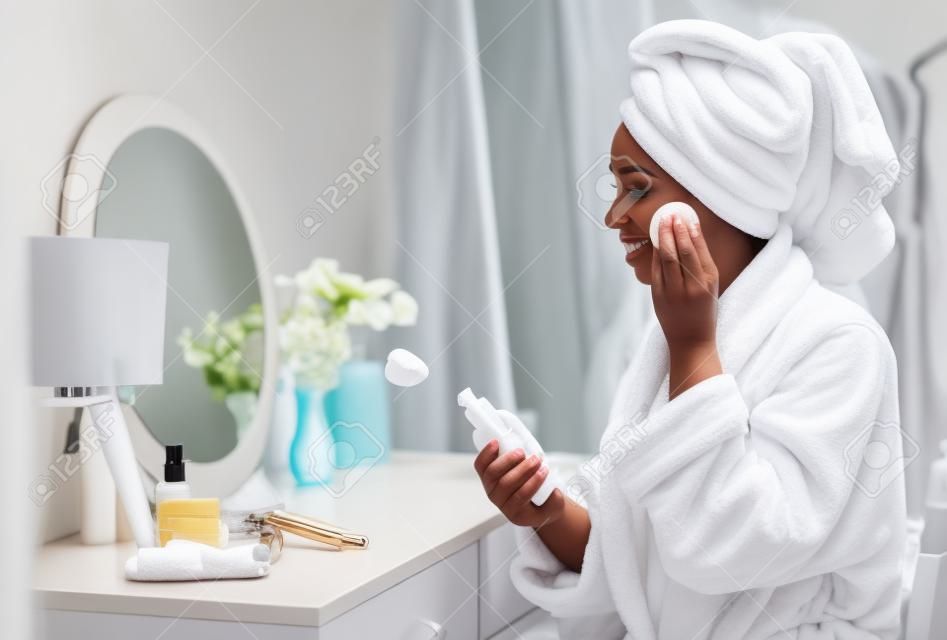Mooie zwarte vrouw schoonmaken gezicht van make-up met reinigende melk en katoenen pad thuis, verpakt in badjas en handdoek, zittend aan dressing tafel