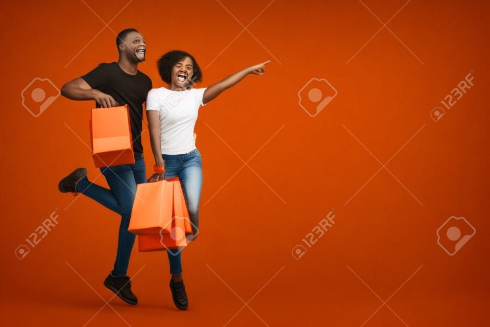 Emocjonalna afro-amerykańska para z zakupami, podskakując i radośnie wskazując na pustą przestrzeń, pomarańczowe tło studyjne. szczęśliwy młody czarny mężczyzna i kobieta dążący do reklamy