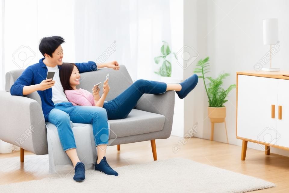 Japanisches Paar, das Handys benutzt, SMS schreibt und im Internet surft, entspannt auf der Couch drinnen sitzt. Freizeit am Wochenende. Glückliche und entspannte asiatische Familie mit mobiler Anwendung, die sich zu Hause ausruht