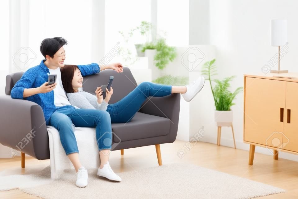 屋内のソファに座ってリラックスして携帯電話のテキストメッセージとブラウジングインターネットを使用して日本のカップル。週末のレジャー。自宅で休んでいるモバイルアプリケーションを使用して幸せでリラックスしたアジアの家族