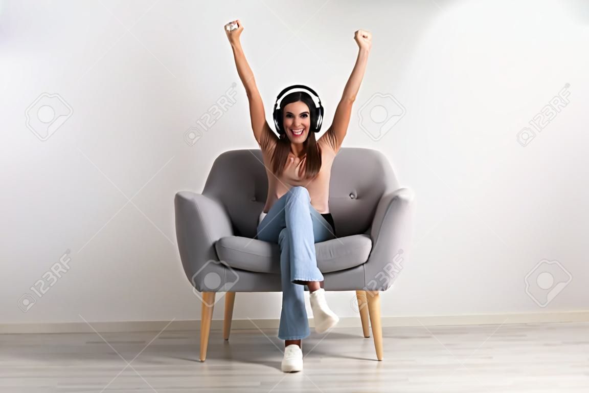 Mujer joven emocionada con auriculares sentada en un sillón, celebrando una victoria en línea, una gran oferta o éxito empresarial