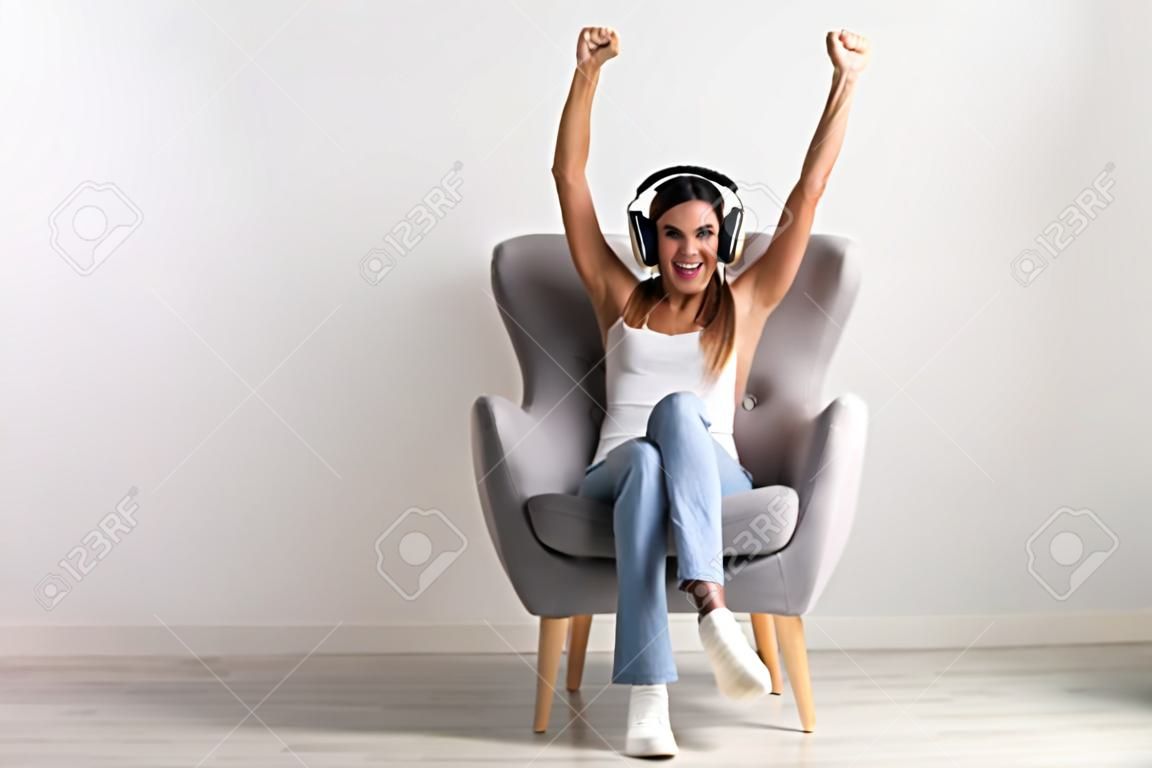 헤드폰을 끼고 안락의자에 앉아 온라인 승리, 큰 거래 또는 사업 성공을 축하하는 흥분한 젊은 여성