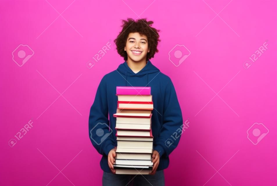 Adolescente alegre de pie con una gran pila de libros, con mucha tarea en el fondo del estudio rosa